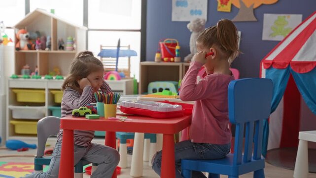 Girl covering ears with hands while friend holds toy stethoscope at small table in preschool building; annoyance play.