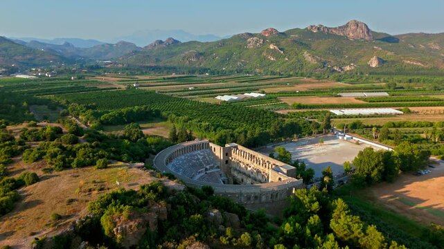 Aerial view of Aspendos Theatre in Antalya. A drone shot shows the ancient Roman amphitheatre surrounded by green hills on a sunny day.