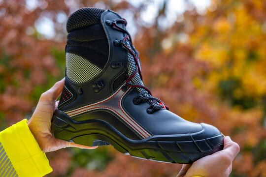 Worker in hi-vis vest holds black safety heavy-duty work boots with prominent reinforced steel toe, protective gear, safety equipment, highest product quality, special shoes for different professions