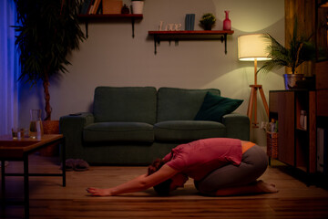 Woman in childs pose on floor, forehead resting, ambient lamp and plant framing scene, slow exhale and surrender to rest evening teacher practicing restorative pause © fotodrobik