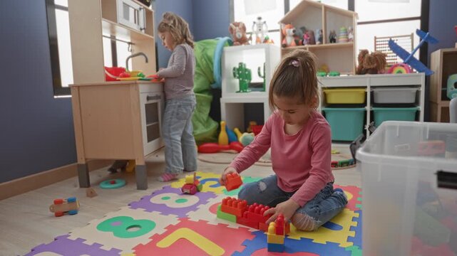 Girl sitting crosslegged on foam mat, hand arranging red building blocks near storage shelves inside a building; concentration creative play.
