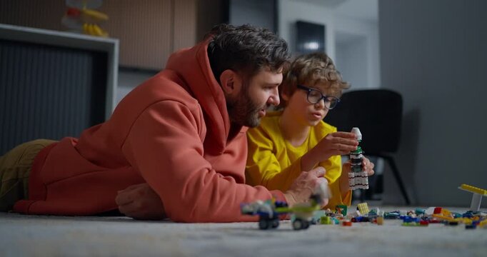 Father and young child lying on living room floor building colorful toy block structures together during playful family bonding at home