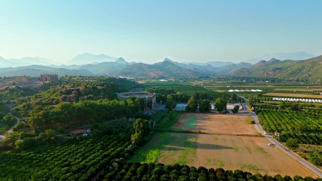 Wide drone view of Aspendos countryside. Ancient ruins and rural landscape stretch toward mountains under clear daylight.
