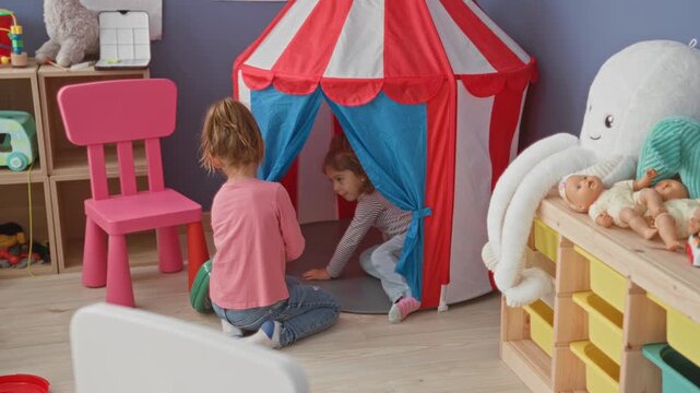 Two girls kneel and reach into a red striped play tent with dolls, one holding a green case inside a preschool building; childhood friendship joy.
