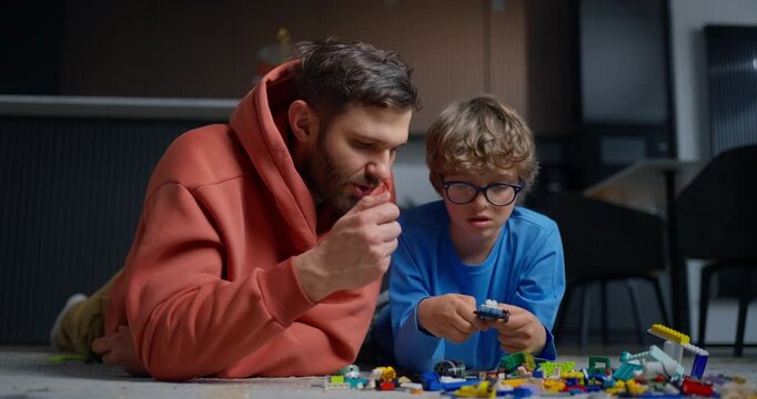 Father and young son lying on the living room floor assembling colorful toy bricks together, sharing a quiet moment of creative play and bonding