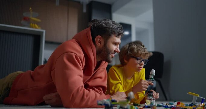 Father and young son lying on floor building colorful toy bricks together, sharing focused family playtime and creative learning at home
