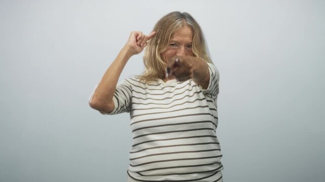 Senior blonde woman points finger directly at camera while gesturing to her temple, wearing striped shirt in studio; assertive challenge.