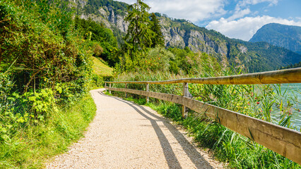 Fototapeta premium Walking path beside a lake with trees and mountains in the background on a sunny day with blue sky and clouds