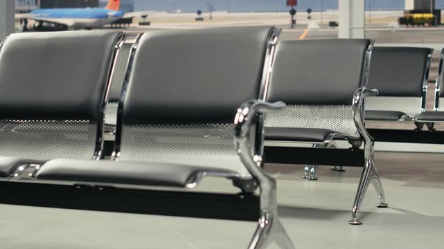 Close up of benches in empty airport hallway following delayed flight schedule. Silent waiting area lounge vacant during unexpected travel disruption, parked plane visible on glass windows