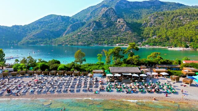 Drone view of Oludeniz Beach, Turkey and mountains. People relax under beach umbrellas by turquoise water on a sunny summer day.