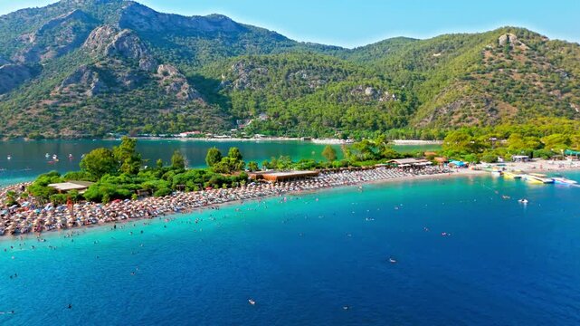 Drone view of Oludeniz Beach, Turkey coastline. People relax under umbrellas along the shore with blue water and mountains in summer.