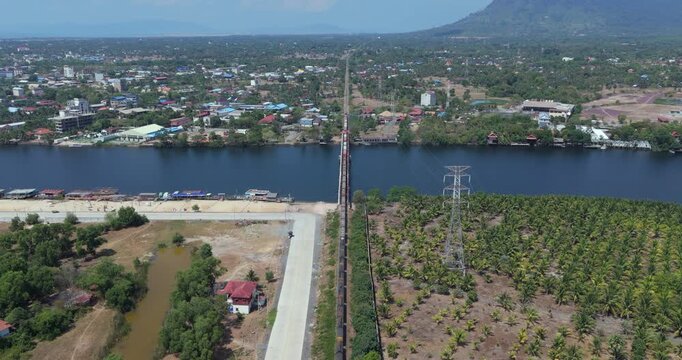 Backward aerial view of a freight train crossing a river bridge in Kampot flanked by a town and palm plantation with distant mountains