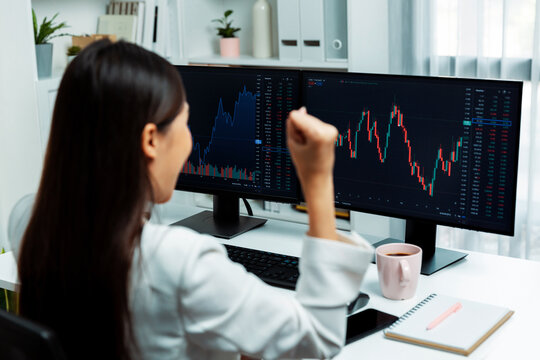 Successful young Asian businesswoman earning highest profit with fist up in exchange stock market on dynamic business graph on pc screens at modern office, analyzing financial technology. Stratagem.