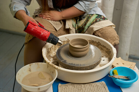 Artisan using a portable heat gun to quickly stiffen a wet clay piece on a spinning pottery wheel.