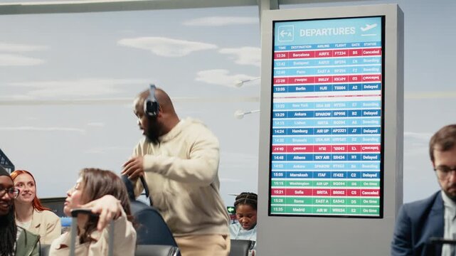 Frustrated tourists watching red status update on screen showing cancelled flights. Concerned travelers holding luggage observing delayed flight announcements on FIDS display, camera A