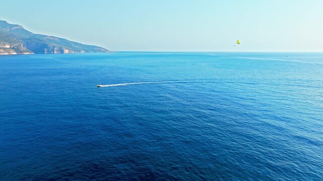 Drone view off Oludeniz Blue Lagoon, Turkey with parasailing. Speedboat pulls a yellow parasail across deep blue sea on a sunny summer day.