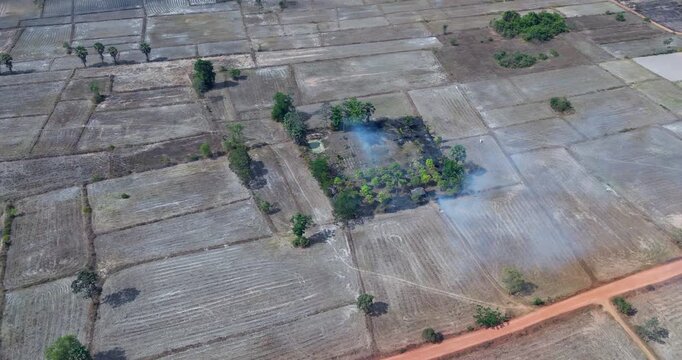 Turrning aerial view of parched, rectangular fields with smoke rising from a green grove. A brush fire chars the earth in the center, while a red dirt road borders the patterned landscape.