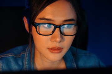 Fototapeta premium Portrait young beautiful Asian creative with glasses of serious face looking on pc to search project creator social media online, planning strategy creator at neon modern office at night. Stratagem.