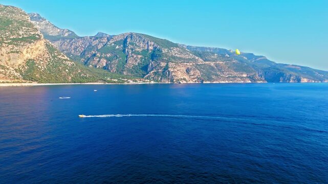 Drone view off Oludeniz Blue Lagoon, Turkey with parasailing. Speedboat pulls a yellow parasail across deep blue sea on a sunny summer day.