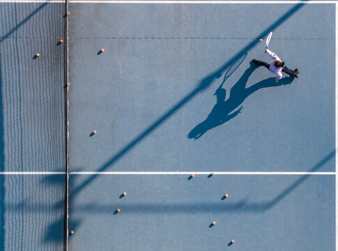 Aerial view of a young girl playing tennis on a blue court, her long shadow naturally mirroring her dynamic swinging motion.