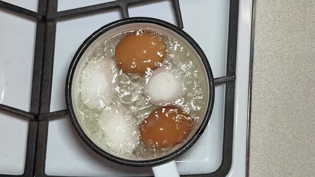 Close-up boiling eggs in enameled ladle on gas stove, brown and white chicken eggs simmering in bubbling water, steam rising, white