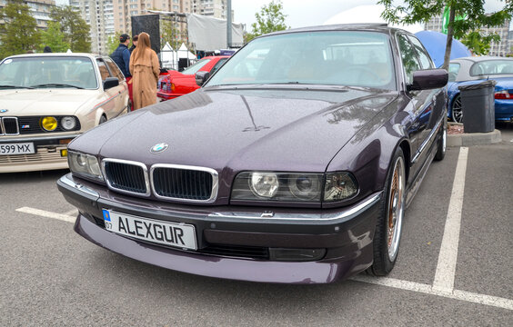 Classic deep purple BMW 7 Series (E38) with elegant wire-spoke wheels. It sits alongside other vintage BMWs at an outdoor car festival. A masterpiece of 90s luxury