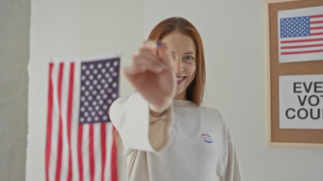 Woman holds tiny american flag pin between thumb and forefinger, hand extended toward camera in a polling building; civic pride.