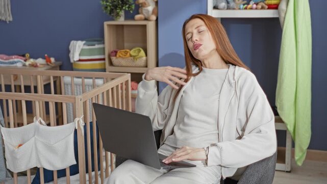 Woman typing on laptop and tucking hair beside wooden crib in a home nursery, adjusting jacket while seated; multitasking fatigue.