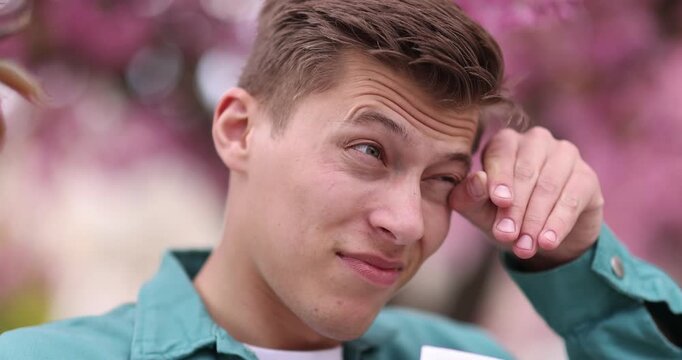 Man with paper tissue suffering from seasonal pollen allergy near blossoming tree on spring day