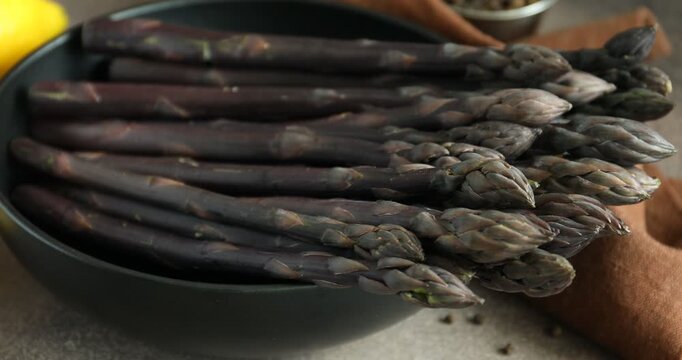 Woman putting raw purple asparagus spears into bowl at grey table, closeup