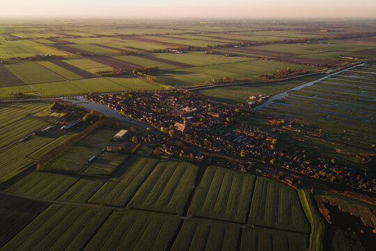 Aerial view of the historic village of Schermerhorn nestled among vast green polder fields and intricate canal networks during golden hour in Schermerhorn, North Holland, Netherlands.