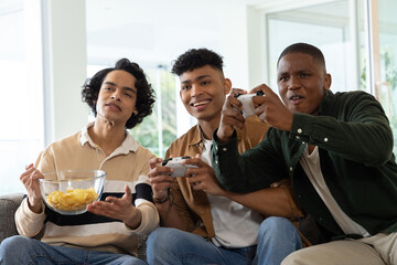 Diverse male friends sitting on gray sofa at home holding white controllers sharing bowl of chips © wavebreak3