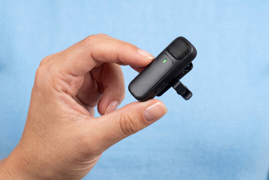 Human hand holding a modern black wireless lavalier microphone against a light blue background