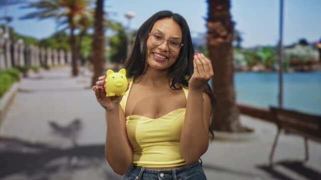 Woman holding yellow piggybank and making coin pinch gesture on a sunlit street promenade near waterfront; savings empowerment optimism.