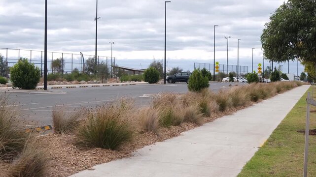 Suburban sports facility car park in Truganina, Melbourne, Victoria, Australia, featuring native tussock grass landscaping with woodchip mulch along a concrete footpath