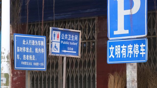Municipal street signs in Chinese and English on an urban street in China, indicating a public toilet 50 metres ahead, a parking area, and a no stopping zone