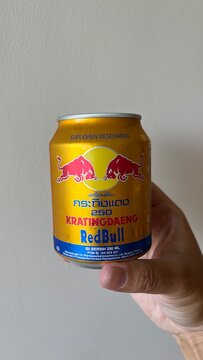 Red Bull Krating Daeng energy drink in golden 250ml aluminum can held by hand against plain wall background indoors - Bali, Indonesia, March 26, 2023