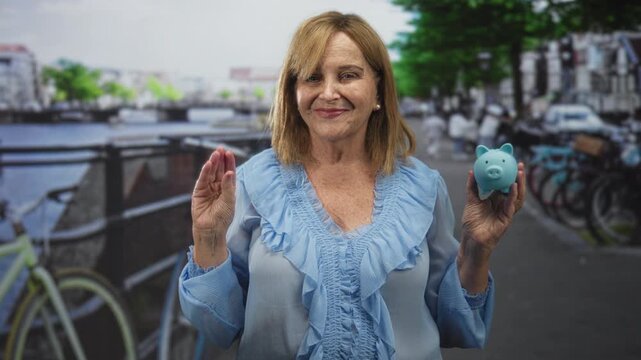 Middle age woman holding a blue piggy bank in her right hand and raising left palm in a pledge gesture on a street by a canal; financial confidence.