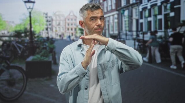 Man makes timeout t gesture with hands on city street, light blue jacket and stubble visible; thoughtful restraint.