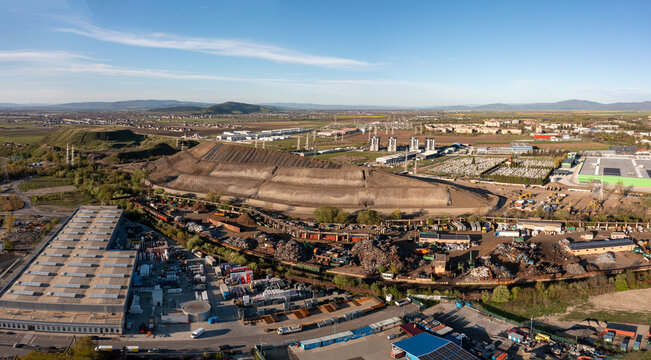 Brasov, Romania - 28 April 2026: Aerial view of an industrial scrap yard and large earthwork mound surrounded by warehouses and a cemetery under a clear sky.