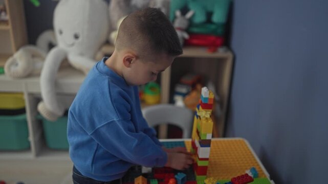 Hispanic boy stacking colorful plastic blocks on a table, small hand grasping a brick inside a playroom in a building; concentration.