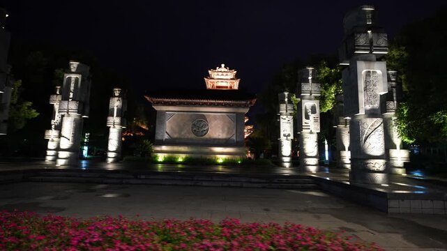 Nighttime illuminated Chinese-style stone carved archway with traditional architectural elements and floral foreground
