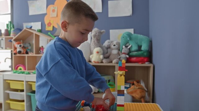 Hispanic boy stacking multicolored blocks into a tall tower with hands in a preschool classroom surrounded by toys and shelves; childhood concentration.