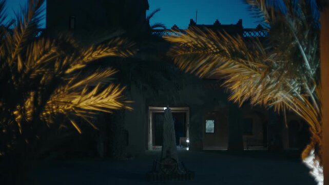 Illuminated palm leaves framing dark clay building with glowing doorway under blue night sky