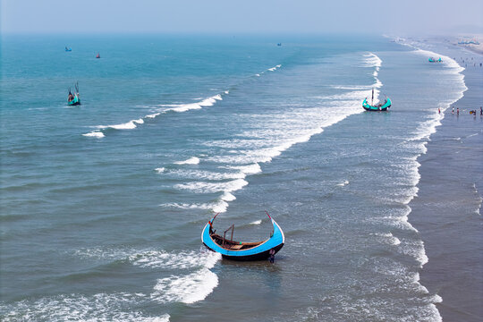 Cox's Bazar, Bangladesh - 10 March 2026: Aerial view of traditional moon-shaped fishing boats called Sampans floating in the turquoise sea and resting on the sandy shore with waves breaking.