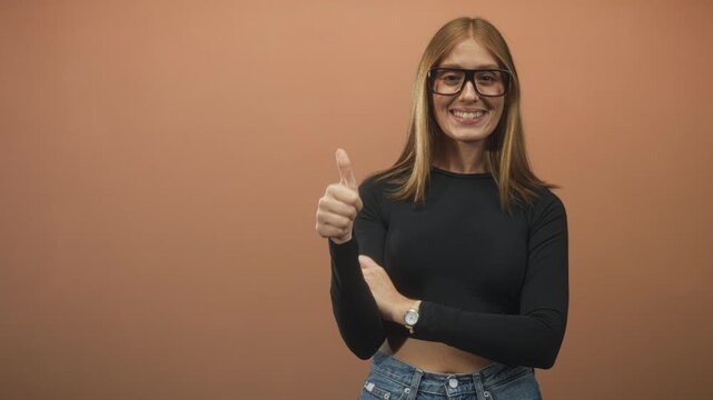 Woman with glasses in black top and jeans shows thumbs up with bare thumb and visible midriff, smiling against a studio backdrop; confidence.