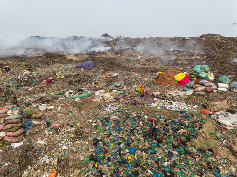 Aminbazar, Bangladesh - 26 January 2025: Aerial view of the Aminbazar landfill with smoke rising from piles of waste and people scavenging through garbage under a hazy sky.