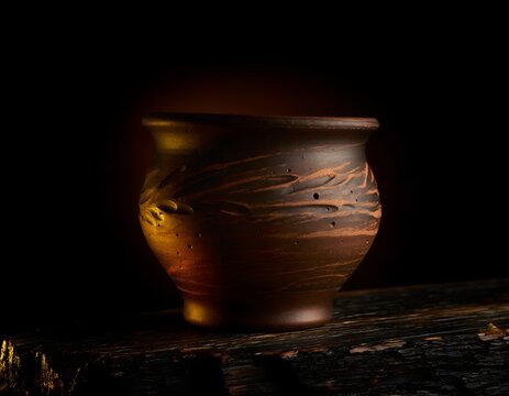 A close up detail shot of a hand thrown unglazed earthenware pot resting on a dark surface highlighting the intricate textured rim and deep earthy tones of the rustic ceramic vessel art object.