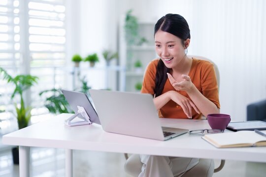 Asian woman hybrid worker video call using laptop computer tablet device smiling happy remote working from modern home office workspace bright natural light