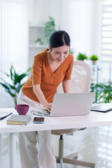 young Asian women working from home office using laptop computer hybrid remote work standing desk bright modern living room interior green plants technology